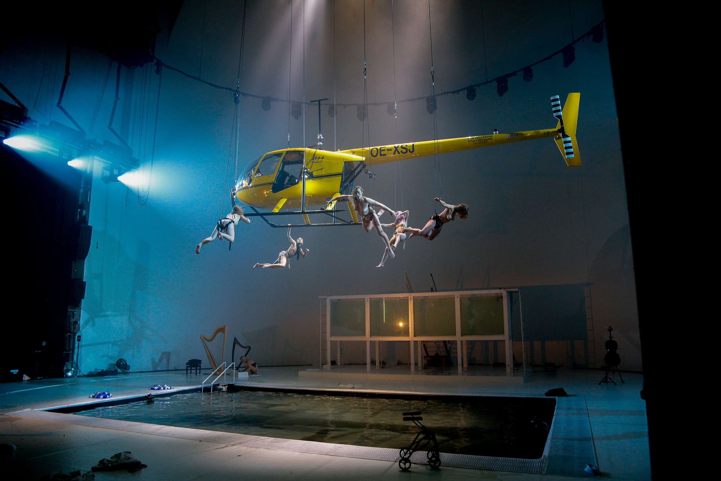 ein Hubschrauber über einer Bühne mit einem Schwimmbecken, an dem mehrere Darstellende hängen
