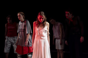 Eine junge Person auf der Bühne in weißem Kleid, dahinter erkennbar das Ensemble