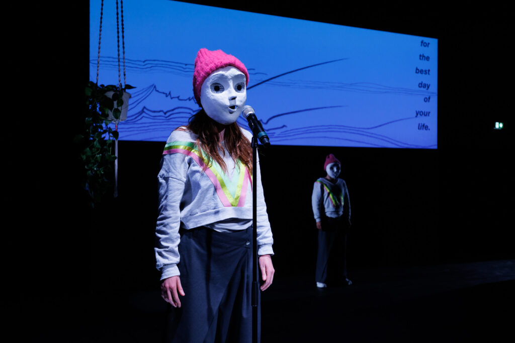 eine Person mit Maske und pinker Mütze vor einem Mikrofon