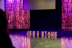 eine Bühne mit pink-lila Glitzervorhang und dem Schriftzug „JES 2 STAGE“